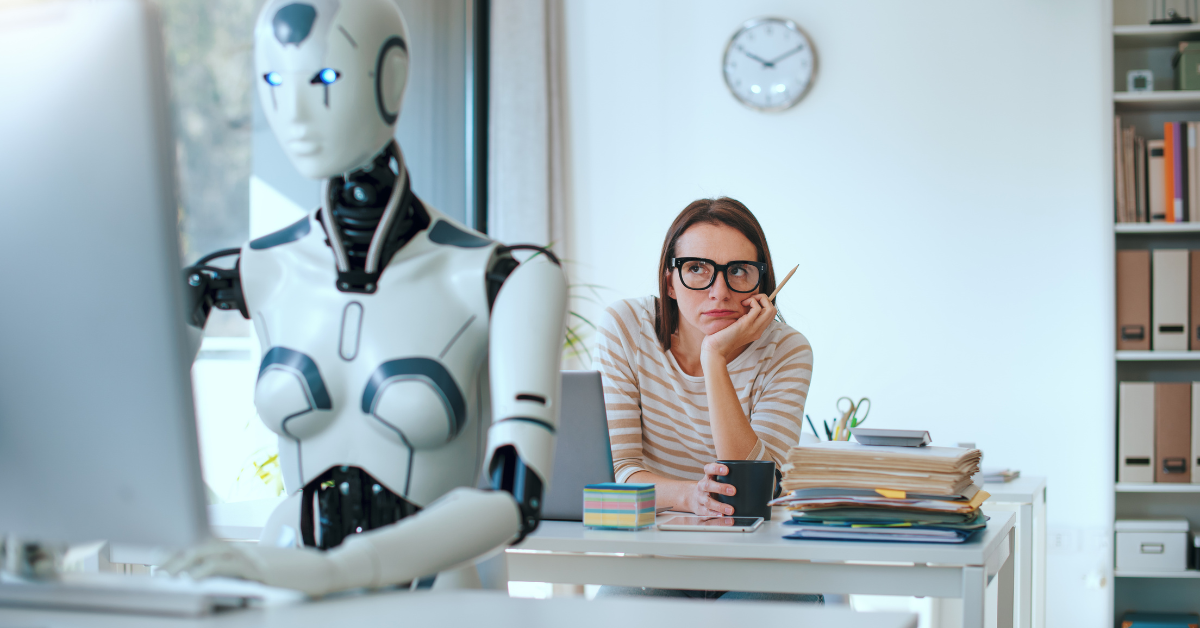 A robot working on a computer as a woman looks over frustrated from her desk.